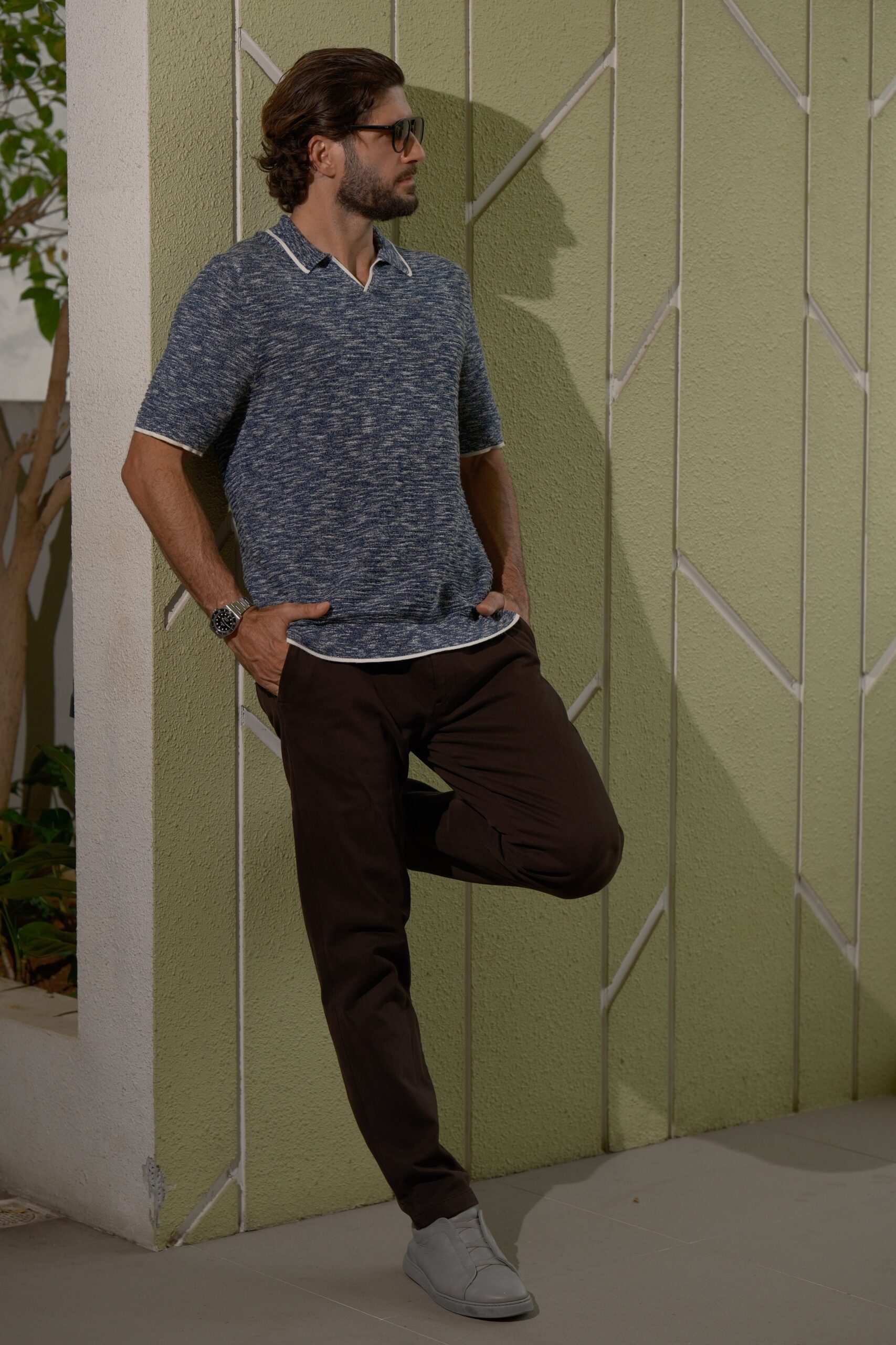 Man wearing a blue marled polo and dark pants leaning against a pale green wall, hands in pockets, looking to the right with sunglasses on.