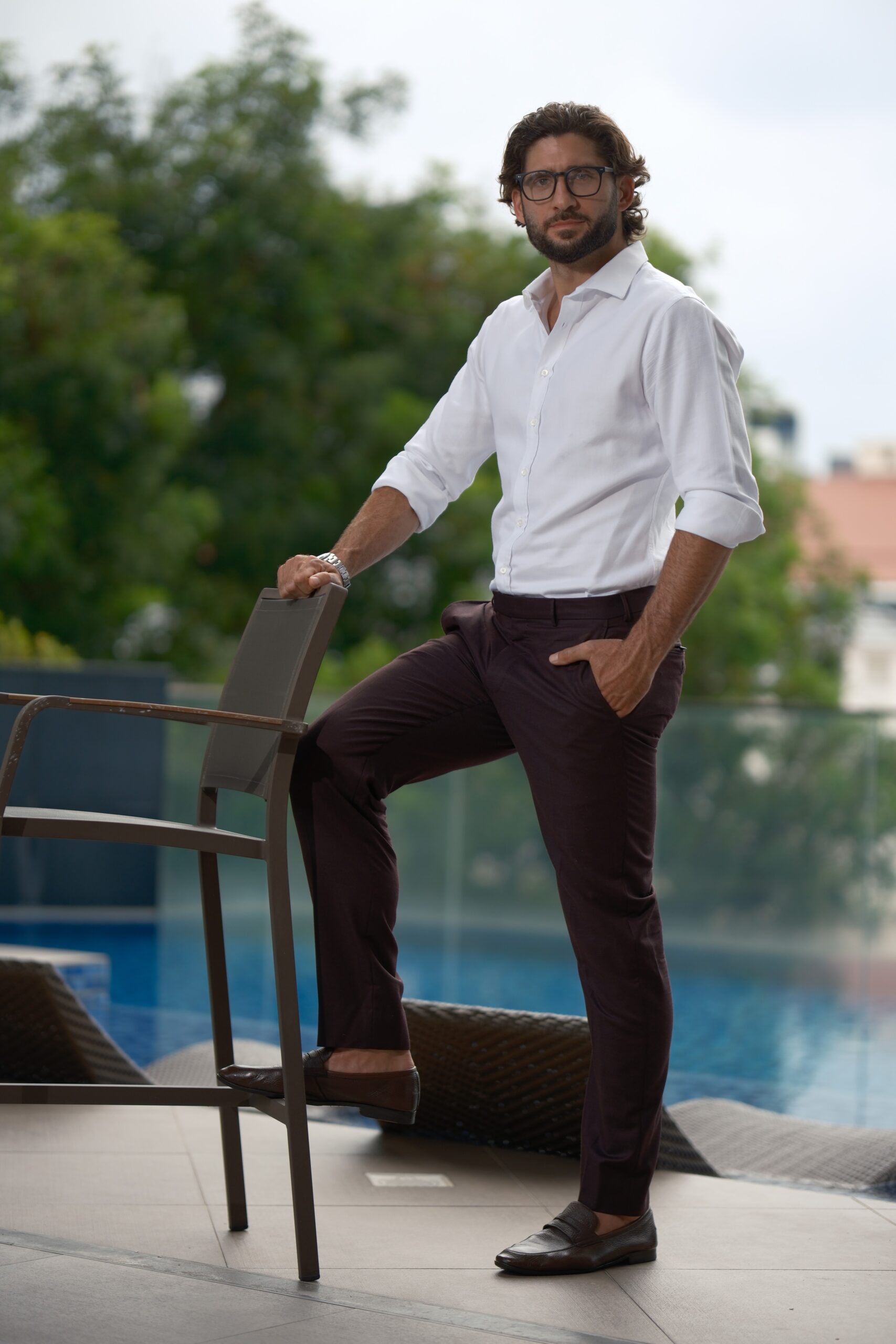 Man in a white button-down shirt and dark trousers leaning on a patio chair beside a pool with green trees in the background, looking at the camera.